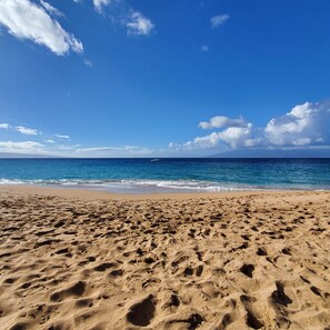 A sandy beach with footprints leading to the water, a clear sky, and a distant sailboat.
