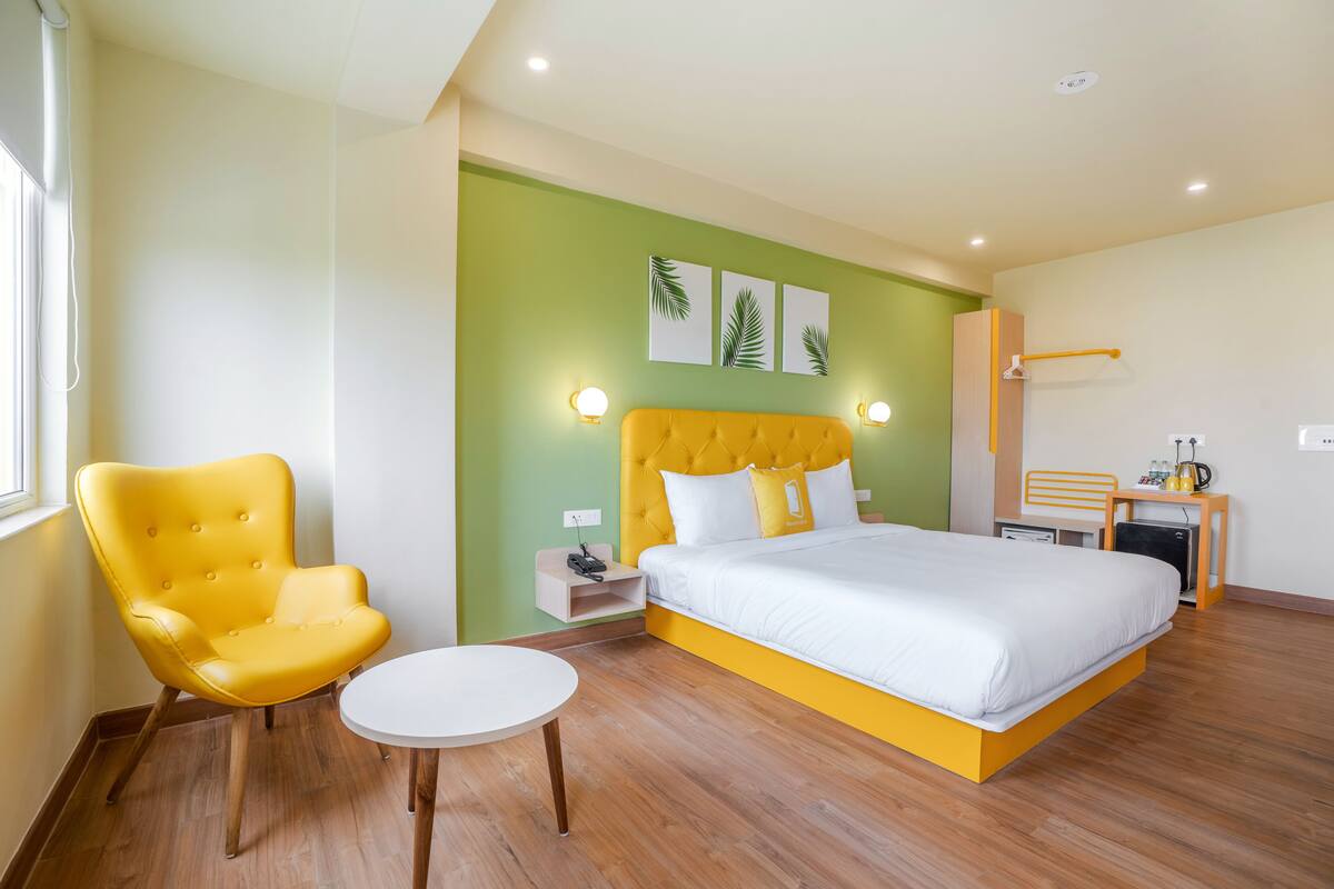 A modern hotel room with a large bed, a yellow armchair, a small round table, and a nightstand with a lamp.