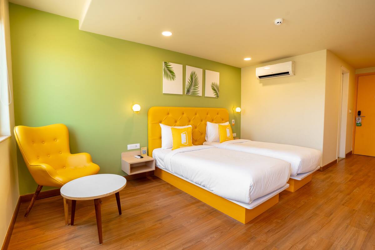 A hotel room with a bright yellow armchair, a bed with white bedding, a round table, and a wall-mounted air conditioner.
