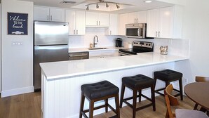 A modern kitchen with white cabinetry, stainless steel appliances, and a central island with bar stools. - The Barefoot Getaway (Stuart)