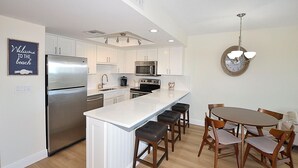 A modern kitchen with white cabinetry, stainless steel appliances, a central island with bar stools, and a dining area with wooden chairs. - The Barefoot Getaway (Stuart)