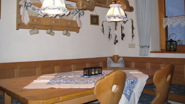 Una mesa de comedor de madera con mantel estampado, sillas de madera y una estantería de madera montada en la pared con luces colgantes y un farol.