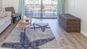 A living room with a sofa, a coffee table, a starfish rug, a wicker ottoman, and a sliding glass door leading to a balcony with a view of buildings.