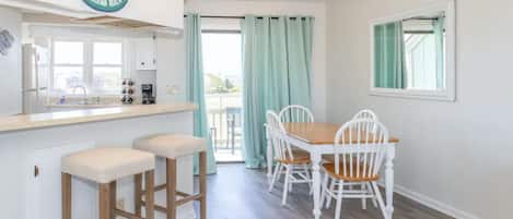 Una cocina con el símbolo CB, un comedor con mesa de madera y sillas blancas, y vistas a través de una puerta de cristal.