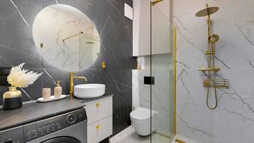 A modern bathroom with a marble backsplash, a round mirror, a gold showerhead, and a washing machine.