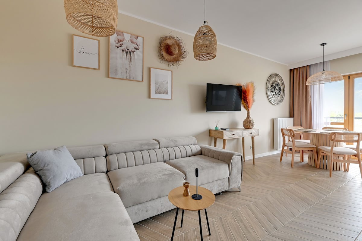 A modern living room with a sectional sofa, a round coffee table, a dining area with a table and chairs, and a television mounted on the wall.