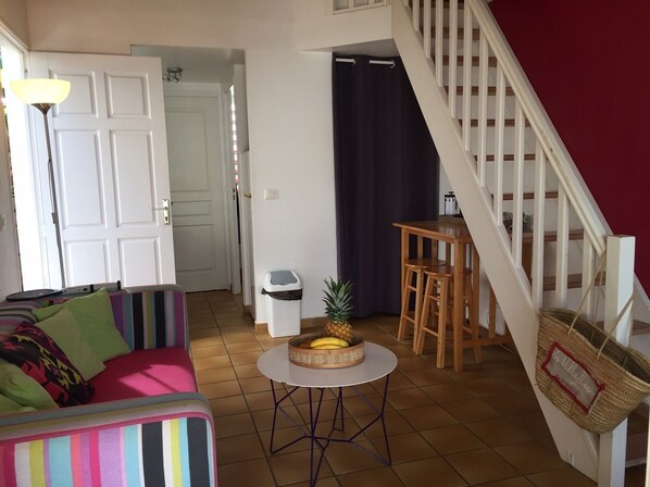 A cozy living room with a colorful sofa, a small round table with a fruit basket, a staircase, and a door leading to another room.