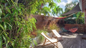 Una terraza de madera con sillones de descanso y una zona sombreada, rodeada de exuberante vegetación y un cerco de madera.