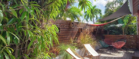 A wooden deck with lounge chairs and a shaded area, surrounded by lush greenery and a wooden fence.