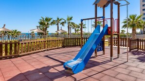 Un parque infantil con un tobogán azul y una estructura para escalar, rodeado de palmeras y una valla de madera.