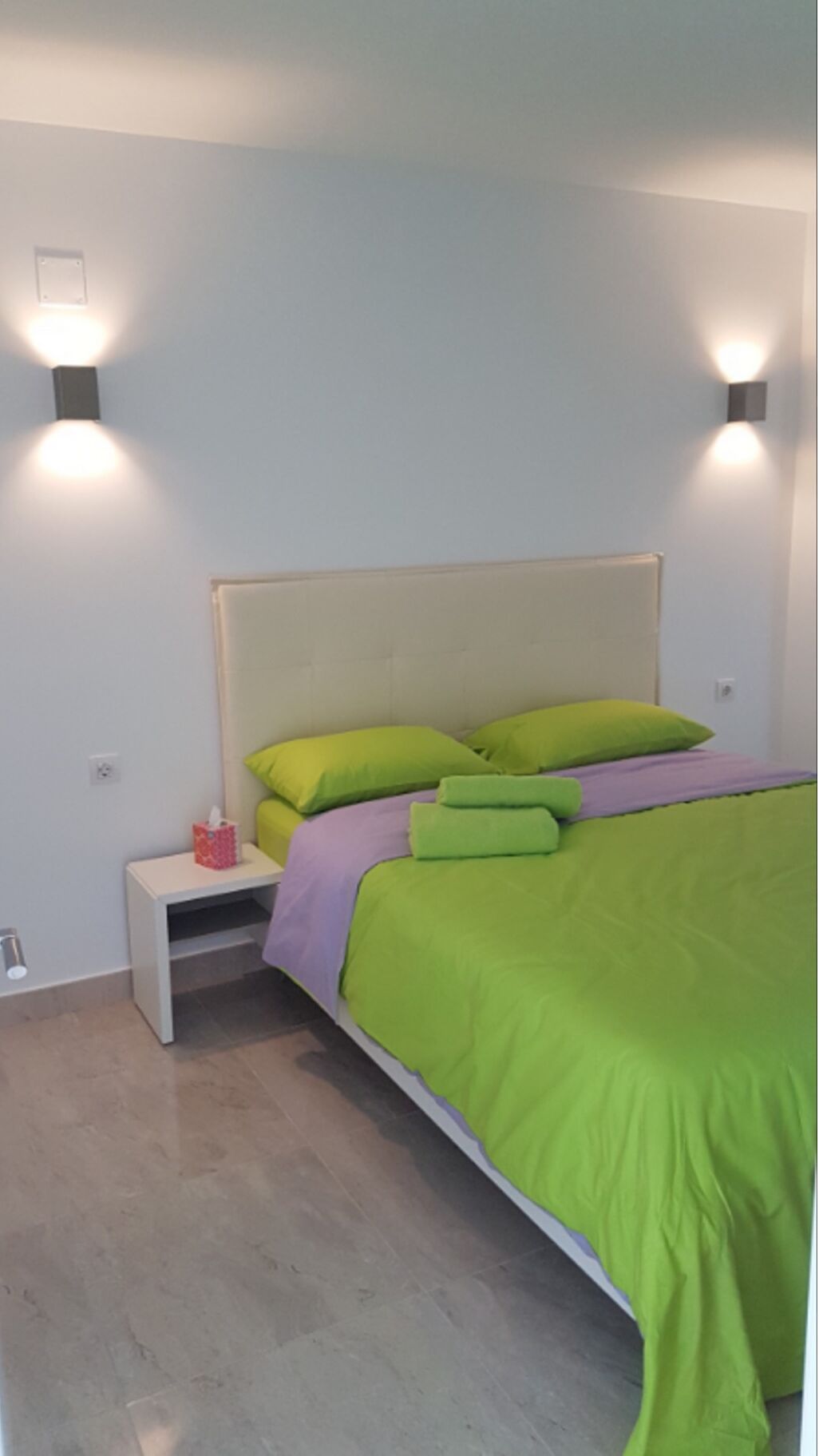 Ein Schlafzimmer mit einem Bett, Bettwäsche in Grün und Violett, einem Nachttisch mit Taschentuchbox und Wandleuchten.
