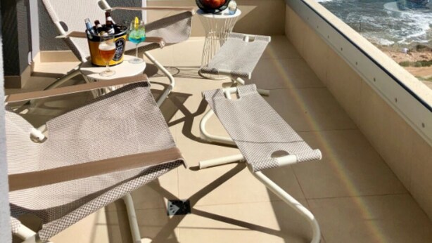 Un balcón con dos sillones, una mesita con frutero y un refrigerador para bebidas. Se ve el océano y un cielo despejado.