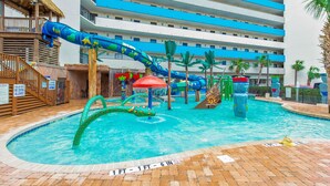 A swimming pool with a water slide, palm trees, and a building with blue and white stripes in the background. - Amazing Direct Modern 2nd Floor Oceanfront w Fireplace (Myrtle Beach)
