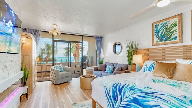 A modern hotel room with a large bed, a balcony with a view of the ocean, a seating area with a sofa and chairs, and a TV mounted on the wall. - Amazing Direct Modern 2nd Floor Oceanfront w Fireplace (Myrtle Beach)
