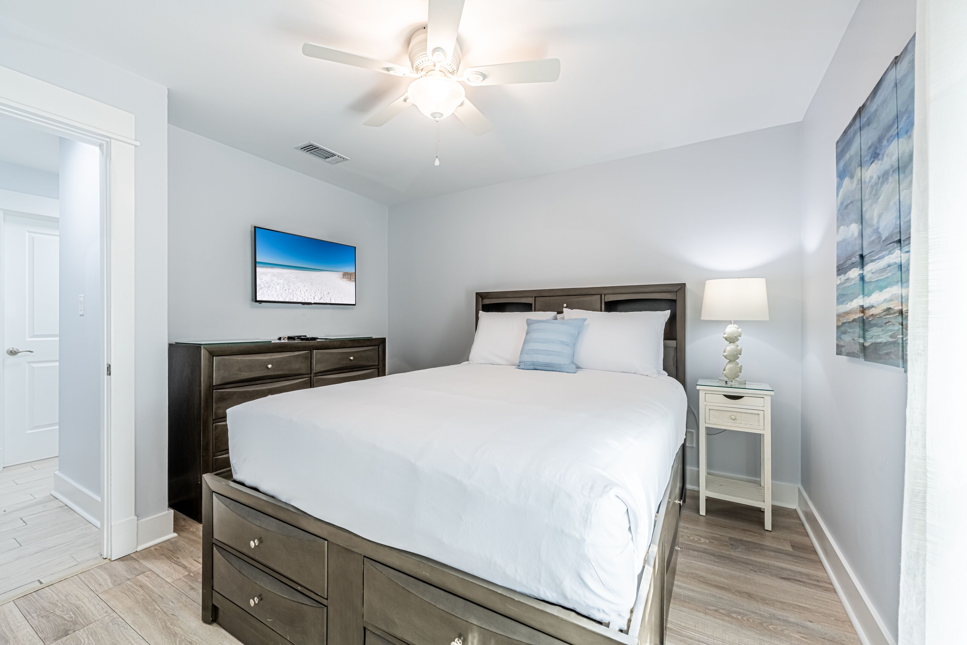 A bedroom with a bed, a nightstand, a ceiling fan, a TV, and artwork on the walls.