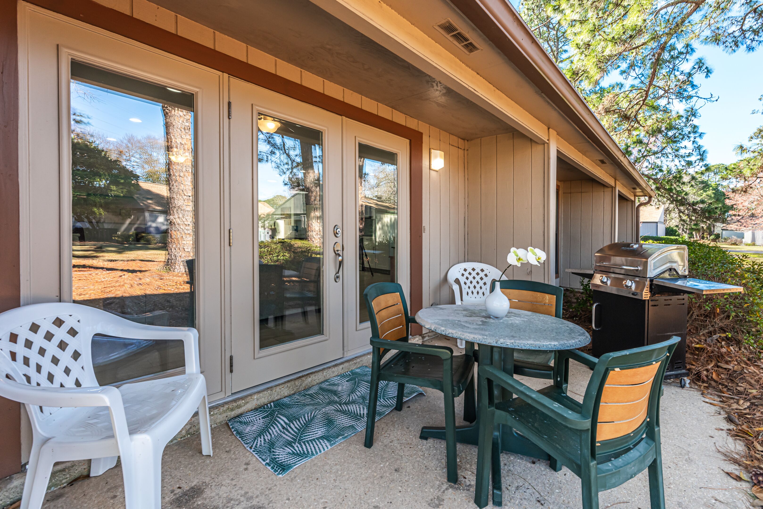An outdoor patio with a round table, chairs, a grill, and a door leading to a backyard.