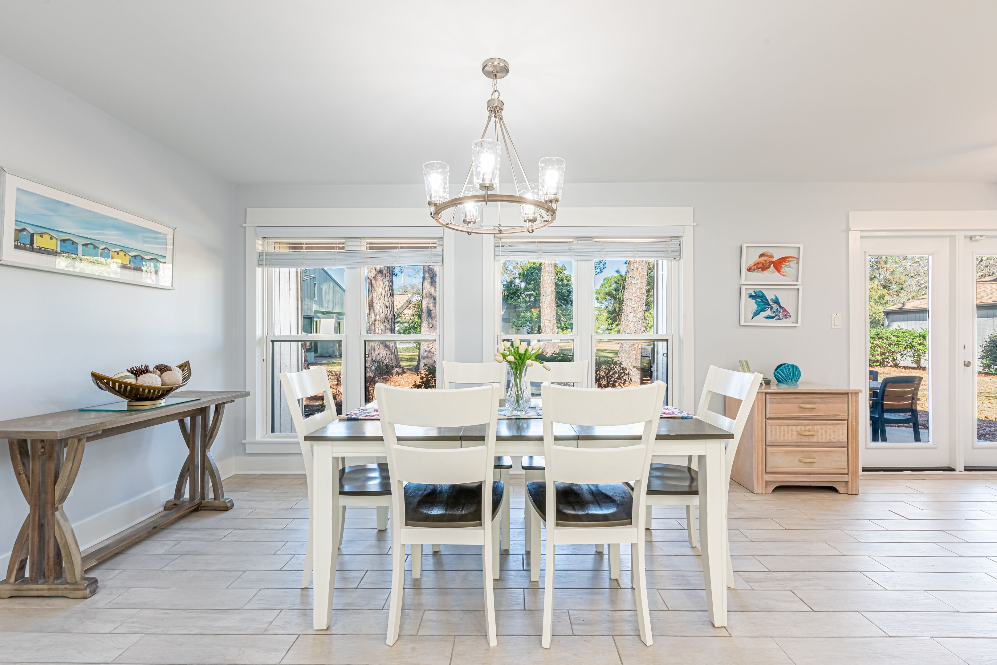 A dining area with a white table and chairs, a chandelier, and large windows with a view of trees.