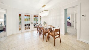 A spacious dining area with a glass table and wooden chairs, a chandelier, and a view through glass doors to an outdoor area. - Sooner Beach Escape: Cute Pool Home in East Rocks (Sanibel)