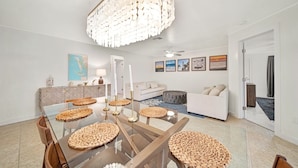 A modern dining area with a glass table, woven placemats, and a chandelier. - Sooner Beach Escape: Cute Pool Home in East Rocks (Sanibel)