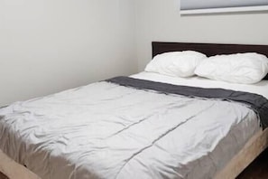Un lit bien fait, avec une literie blanche et une tête de lit sombre, dans une pièce simple aux murs blancs.