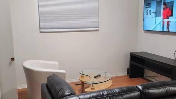 Een woonkamer met een zwarte leren bank, een glazen salontafel, een witte fauteuil en een aan de muur bevestigde televisie.