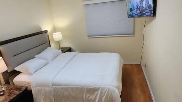 A neatly made bed in a hotel room with a headboard, a bedside table with a lamp, and a television mounted on the wall. - Chesskings Guest House - Unit 2 Notredame Avenue (Winnipeg)