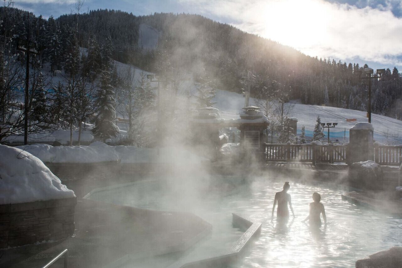 A hot spring area with steam rising, surrounded by snow-covered trees and a mountainous backdrop.