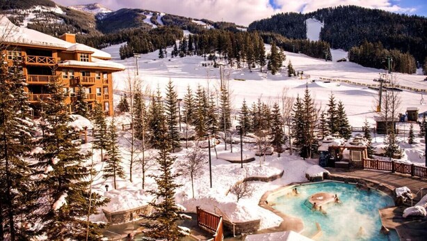 Un resort con una vasca idromassaggio circondato da alberi innevati e con le montagne sullo sfondo.