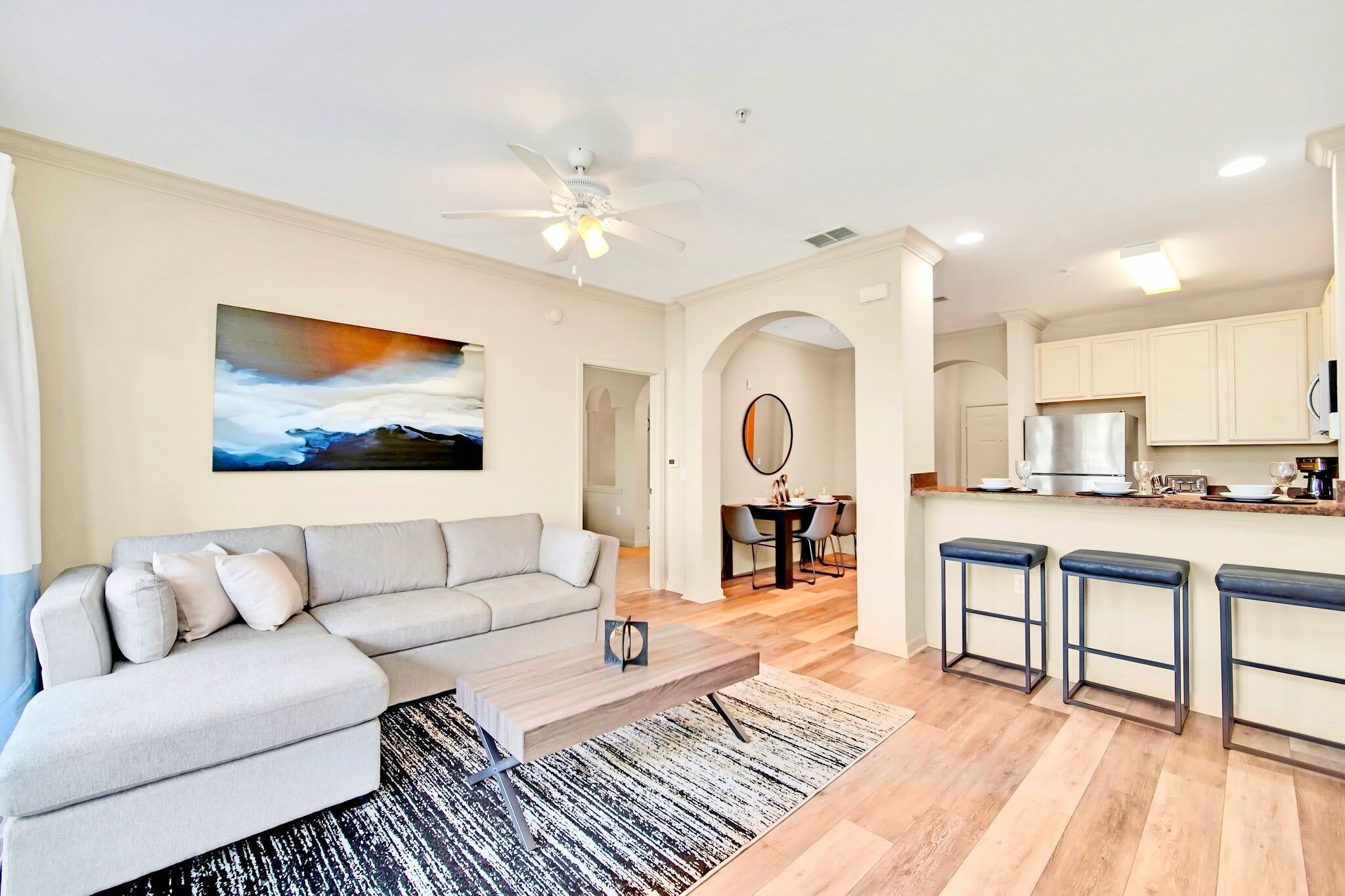 A modern living room with a sectional sofa, a coffee table, and a painting on the wall. There is a kitchen area with a refrigerator, a microwave, and a dining table with chairs.
