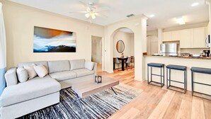 A modern living room with a sectional sofa, a coffee table, and a painting on the wall. There is a kitchen area with a refrigerator, a microwave, and a dining table with chairs. - Upstay 3BR Suite w Pool, Sauna & Games—Near Disney (Winter Garden)