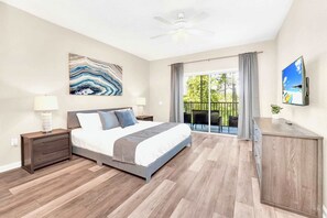 Une chambre à coucher comprenant un lit, des tables de chevet, un ventilateur de plafond, un tableau, un téléviseur et un balcon aménagé avec du mobilier d’extérieur.
