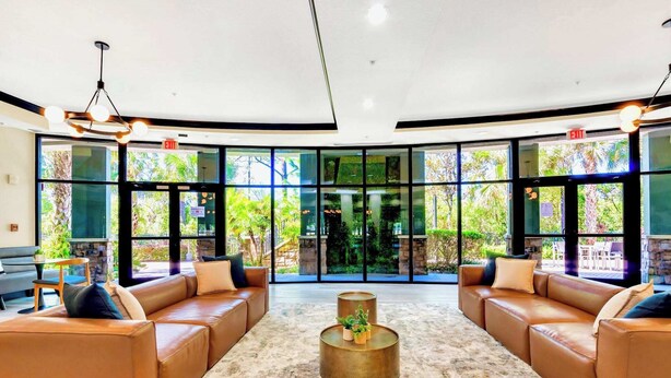 A spacious lobby with leather sofas, a central coffee table, and large windows offering a view of greenery. - Near Disney - Family Condo w Pool, Games, and More (Winter Garden)