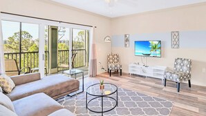 A spacious living room with a sofa, a coffee table, and a TV. There is a balcony with a view of trees and buildings. - Near Disney - Family Condo w Pool, Games, and More (Winter Garden)