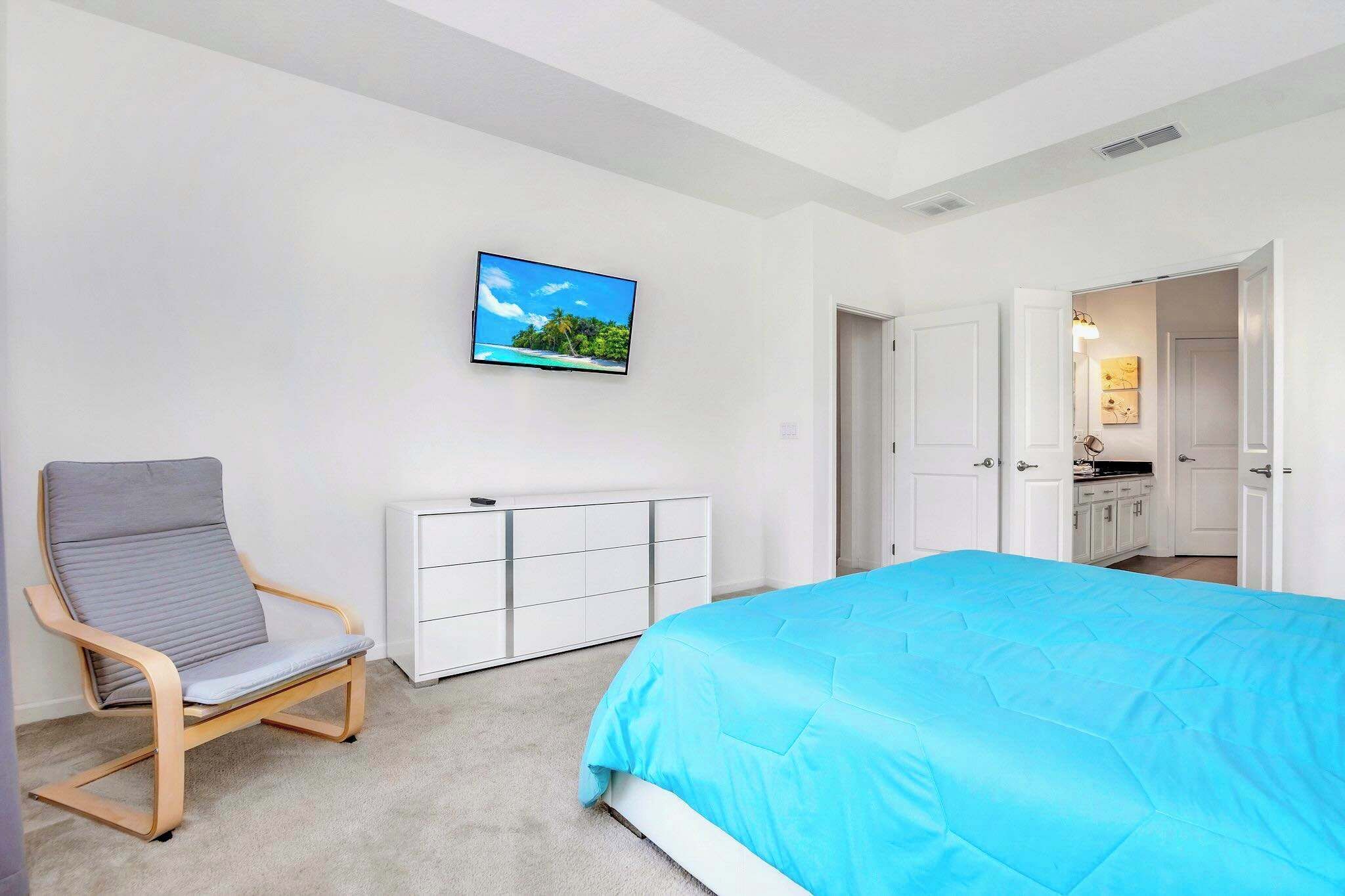 A bedroom with a bed, a chair, a TV, a white wardrobe, and a door leading to a bathroom.