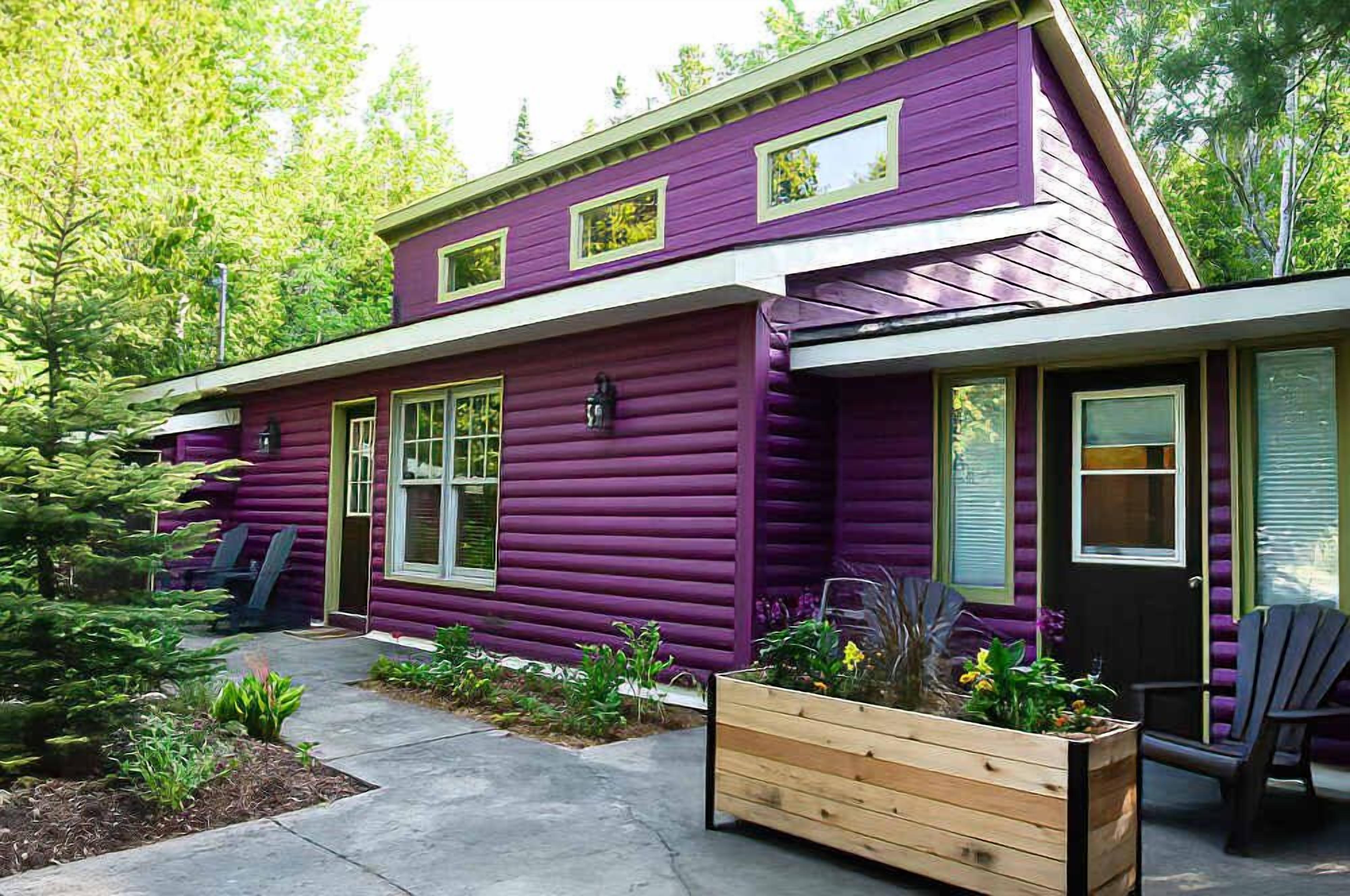Une maison à deux étages avec un revêtement extérieur violet, un porche à l’avant avec des chaises et un jardinier en bois rempli de plantes.