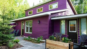 Une maison à deux étages avec un revêtement extérieur violet, un porche à l’avant avec des chaises et un jardinier en bois rempli de plantes.