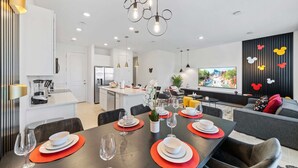A modern living room with a large flat-screen TV, a grey sofa with red cushions, and a dining area with a dark wooden table set for four.