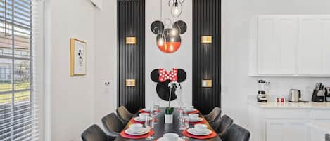A dining area with a long table set for six, flanked by black chairs, with a Mickey Mouse themed light fixture above.