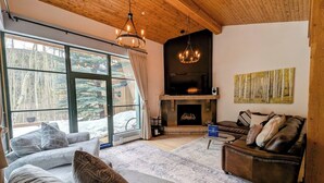 A cozy living room with a fireplace, large windows, and a wooden ceiling.