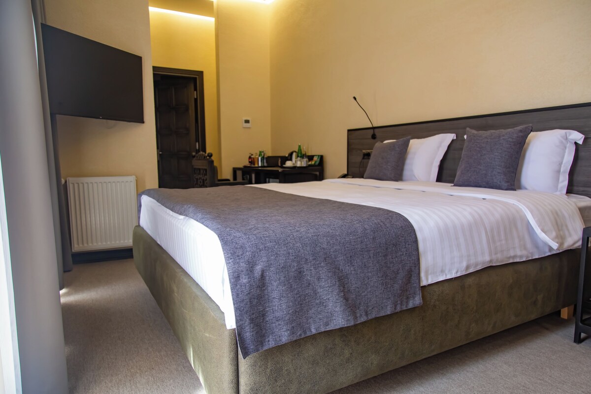 Quarto com cama, mesa de cabeceira, abajur e porta que dá para outro cômodo.