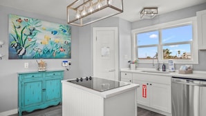 Une cuisine moderne avec un îlot central, des meubles blancs, un meuble de couleur turquoise, un tableau représentant des poissons et une fenêtre donnant sur des palmiers.