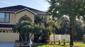 Una casa de dos pisos con fachada color beige, un garaje y un pequeño jardín delantero con palmeras y plantas en macetas.