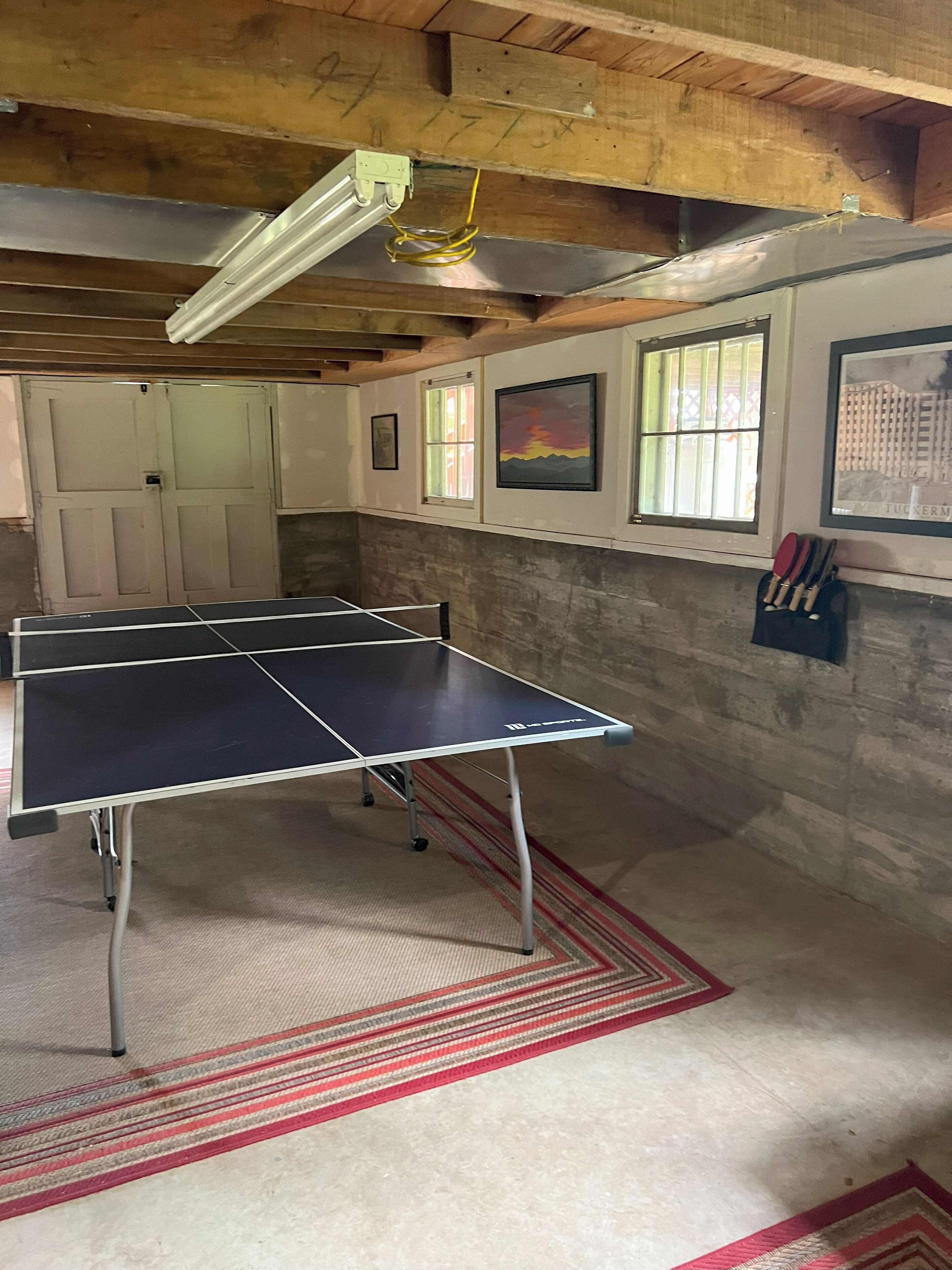 A ping pong table in a room with wooden beams, a window with bars, and a framed picture on the wall.