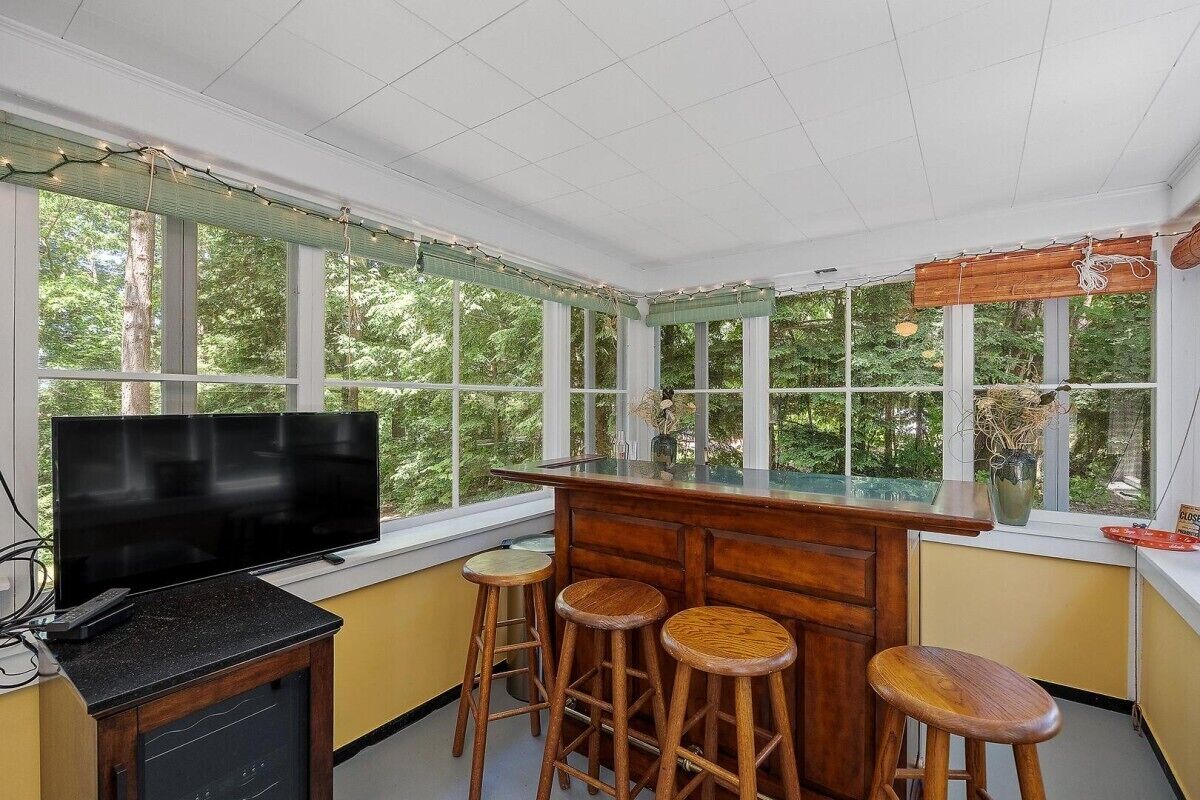 Un solárium con televisor, taburetes de madera y ventanas amplias con vistas a árboles.