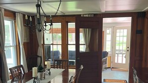 A dining room with a wooden table, chairs, and a chandelier. There is a view of the outside through large windows and a door leading to another room. - Vintage & Modern Lodge-Lake Winnipesaukee water access w/ boat slip, dog welcome (Gilford)