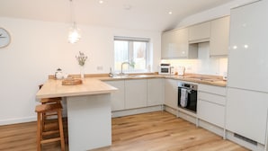 A modern kitchen with wooden flooring, white cabinetry, a central island with stools, a hanging light fixture, and a window providing natural light.