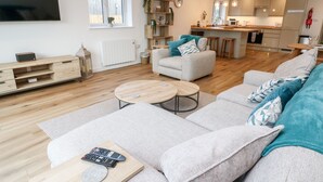 A modern living room with a sofa, two coffee tables, and a TV. There is a kitchen in the background with a dining area.