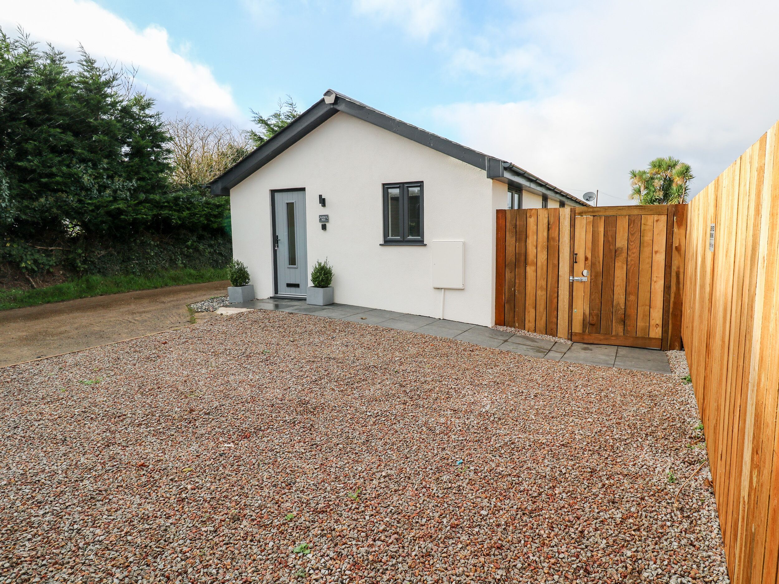 A modern single-story house with a gravel driveway, a wooden gate, and a small front yard with two potted plants.