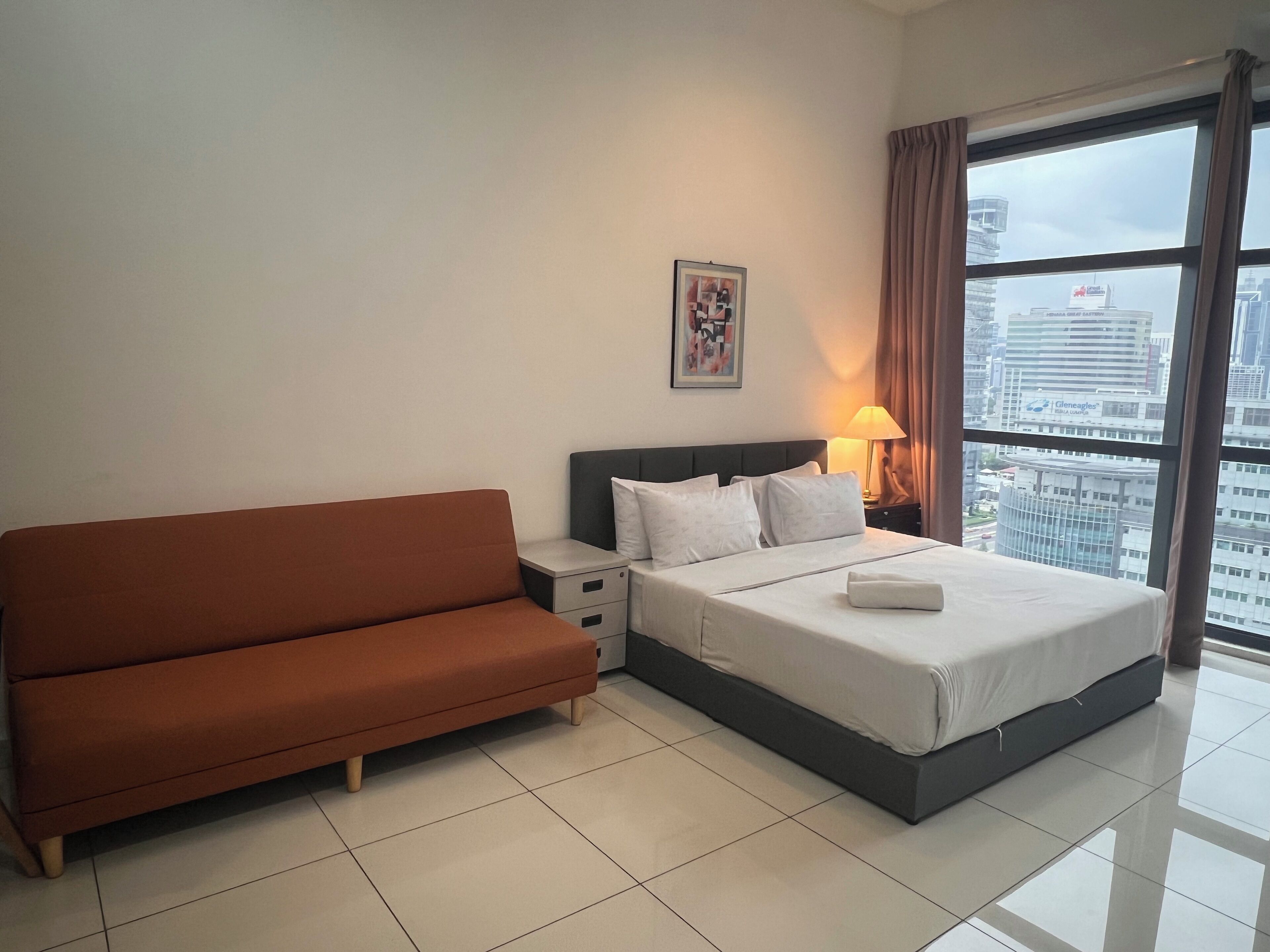 Habitación de hotel con cama, sofá, mesita de noche, lámpara y ventana con vista a la ciudad.