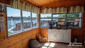 Una baita con pareti in legno e ampie finestre con vista su un lago. Si notano cesti di vimini, un armadio e un cartello con la scritta 'Rilassati! Sei nel tempo del lago'.